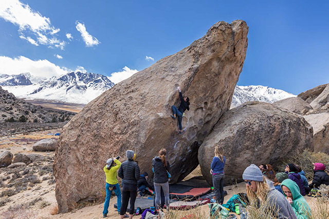 A group of people watch as a climber maneuvers up a boulder. 