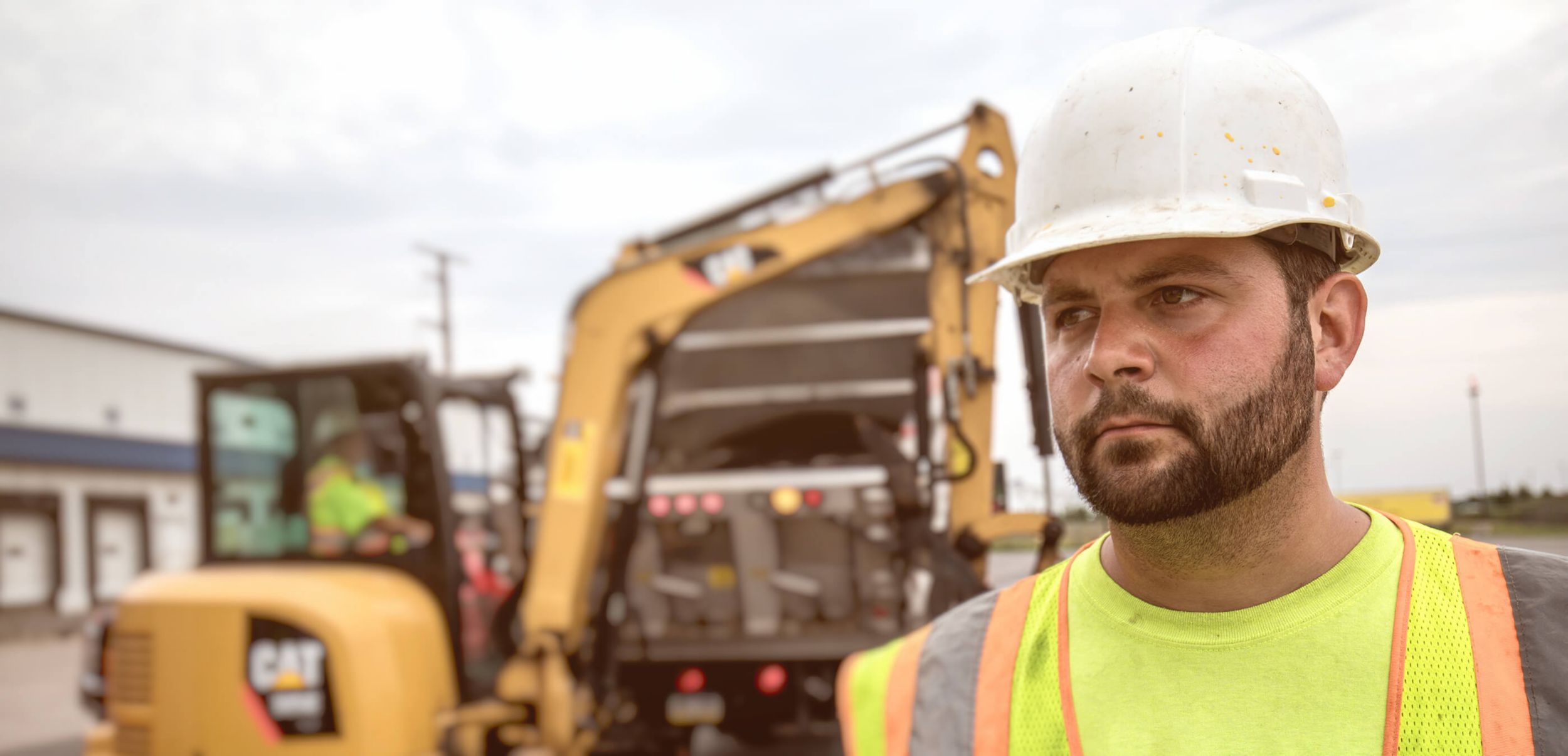 Younger male customer on a jobsite facing forward with a mini excavator working in the background