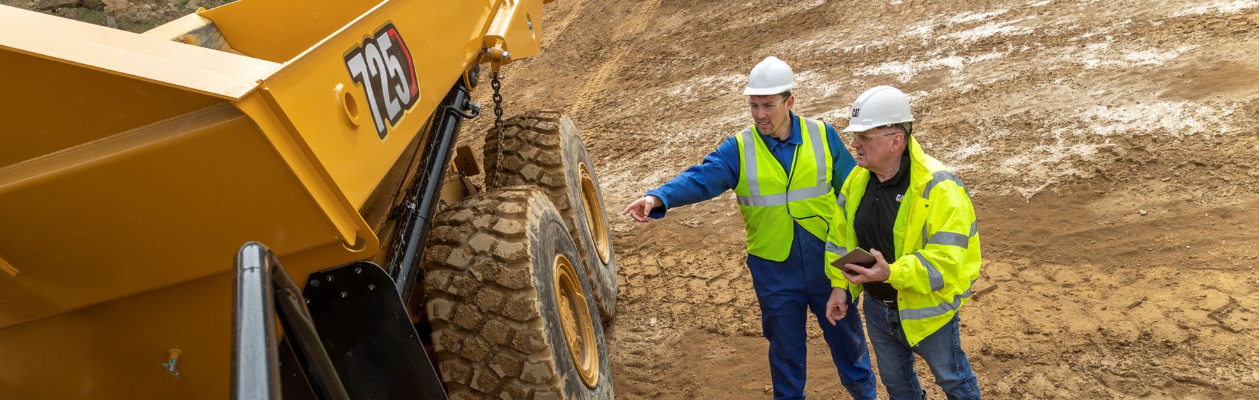 Deux ouvriers portant des gilets à haute visibilité et des casques de sécurité regardent un camion articulé 725 sur un chantier.