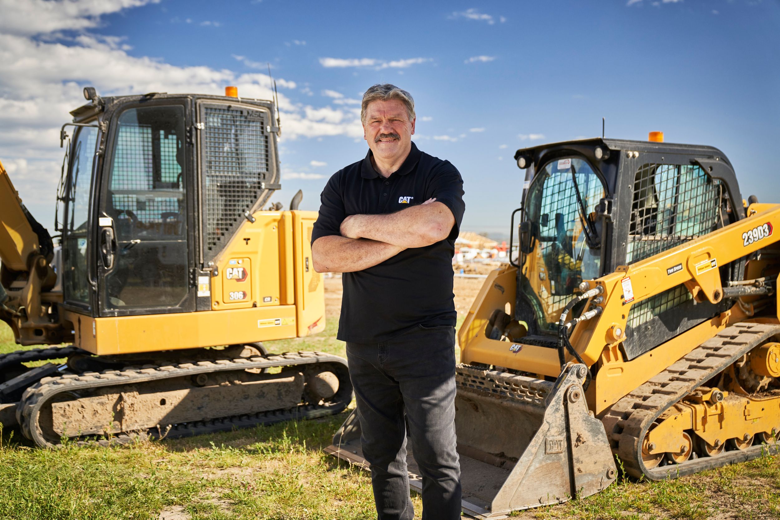 Briak Taylor (BT) standing with his arms folded, in front of two Cat® Compact machines