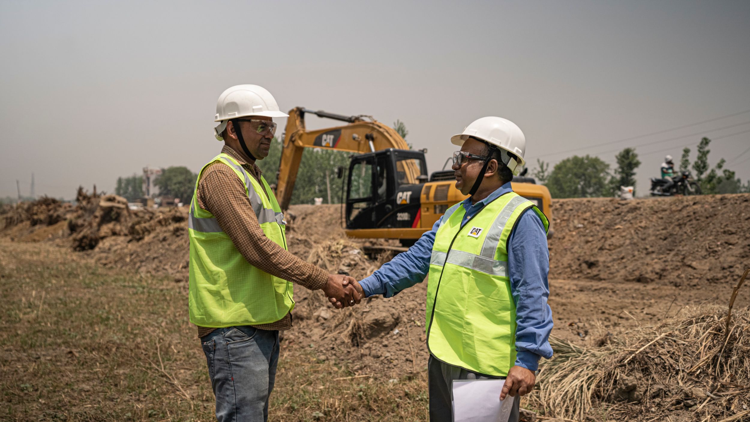 Two Cat® technicians shaking hands out on a bare jobsite in the foreground, with a Cat machine in the background