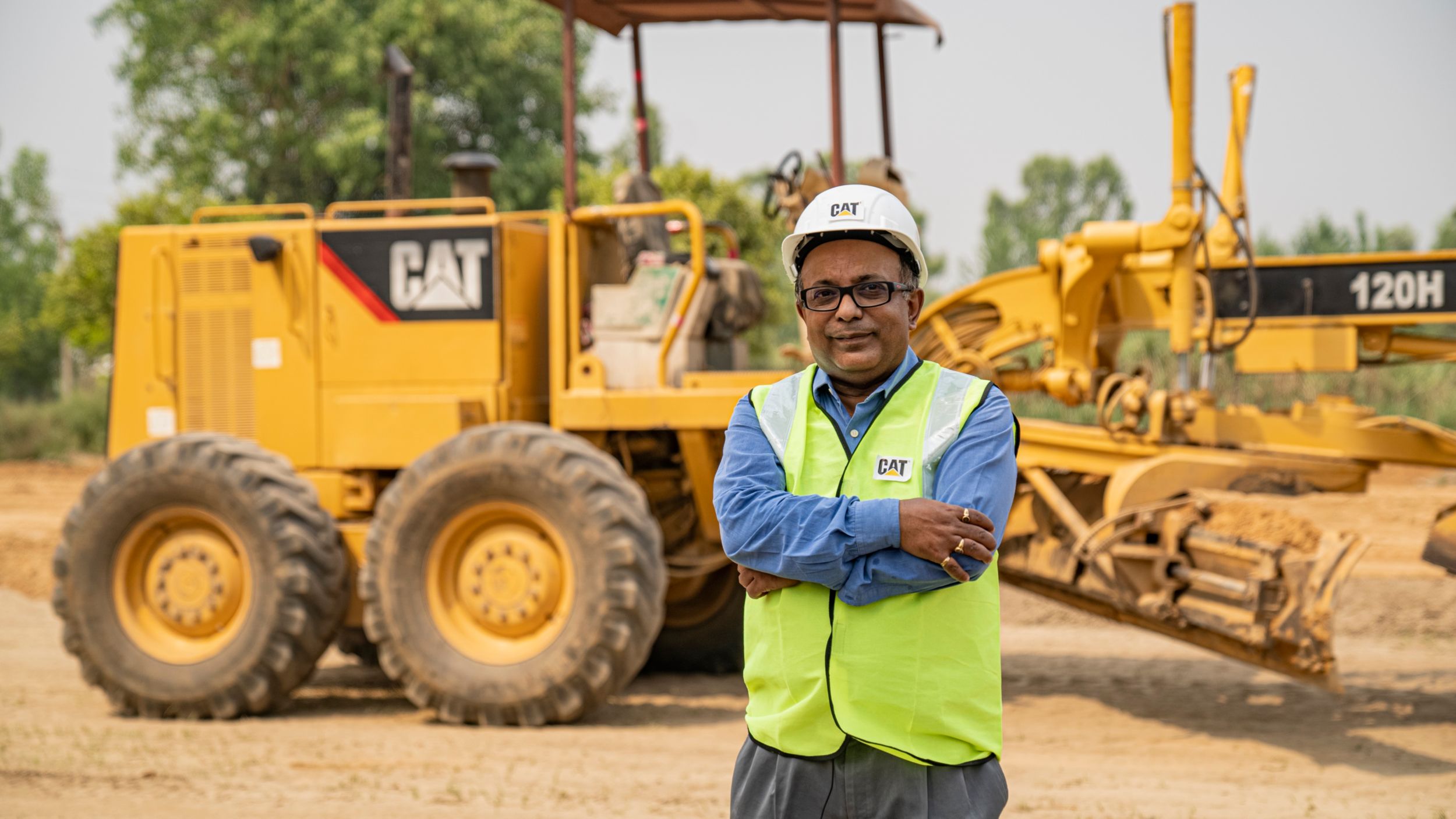 Cat® technician standing with his arms crossed, wearing safety gear, in front of a stationary Cat machine