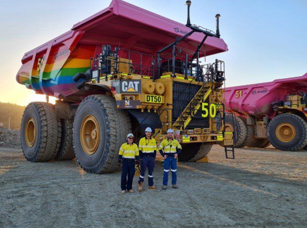 Three people stand in front of a rainbow-colored Cat mining truck