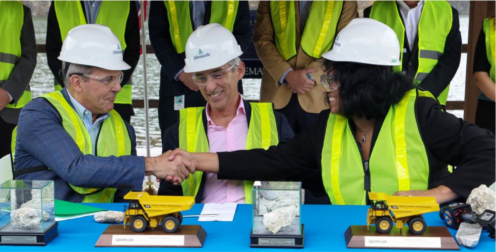 Caterpillar and Albermarle employees shake hands while sitting at a table featuring scale model Cat machines and rocks in display cases
