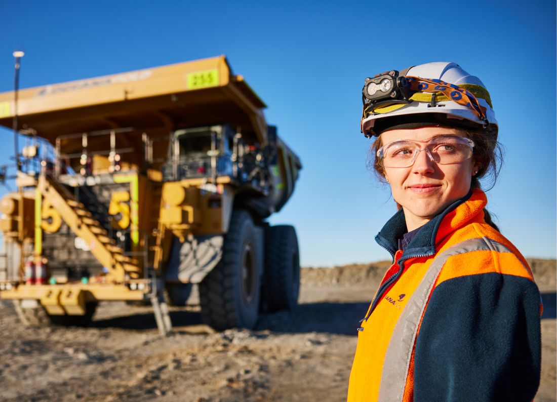 Woman wearing a hard hat and orange jacket while standing in front of a Cat mining truck