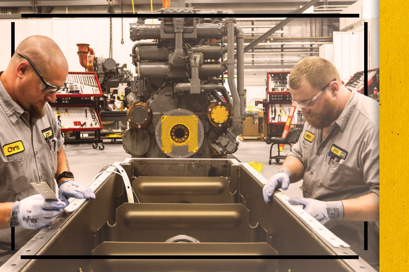Two Cat workers in PPE repairing an engine part with a large engine in the background