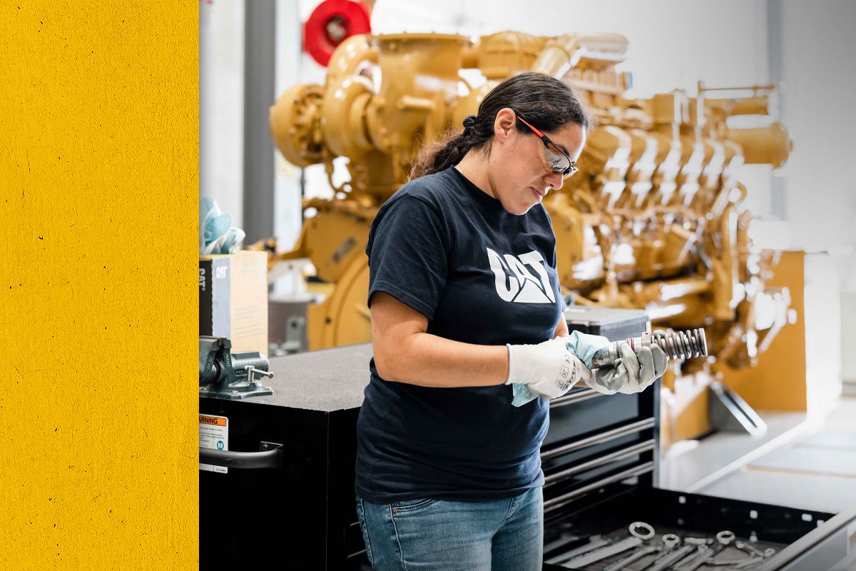A Cat worker in PPE cleaning an engine part with a large engine in the background