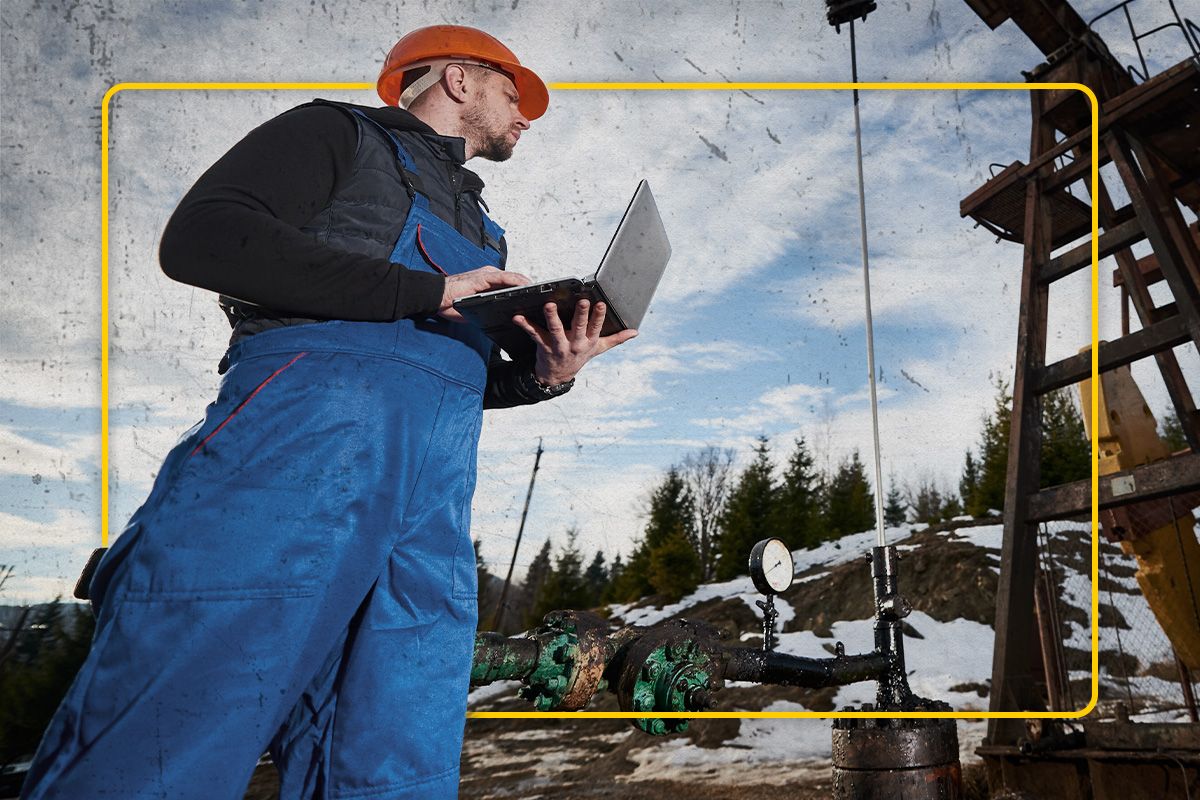Ground up angled view of a Cat worker in coveralls holding a laptop and looking at an oil rig with trees in the background