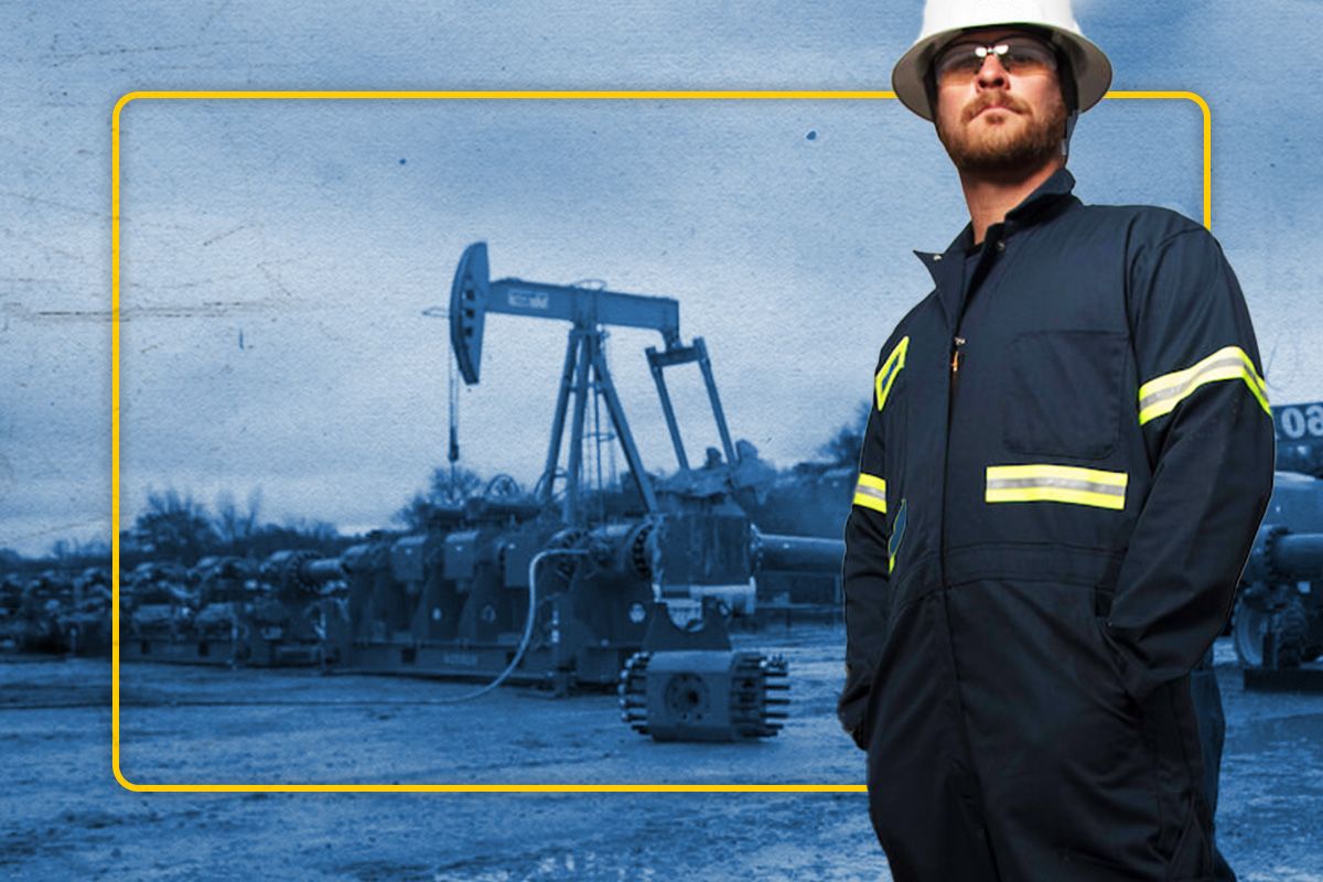 Cat worker in PPE with hands in his pocket looking at the camera with an oil derrick and other machinery in the background