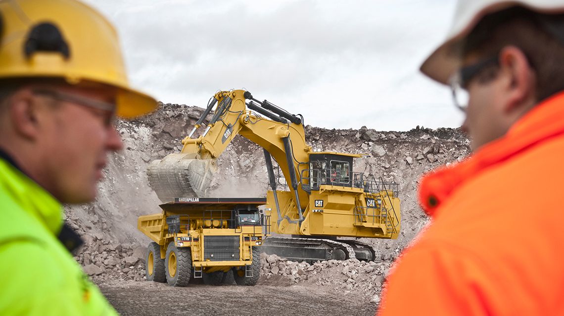dois homens diante de uma mina com uma escavadeira hidráulica de mineração Cat e um caminhão de mineração Cat no fundo