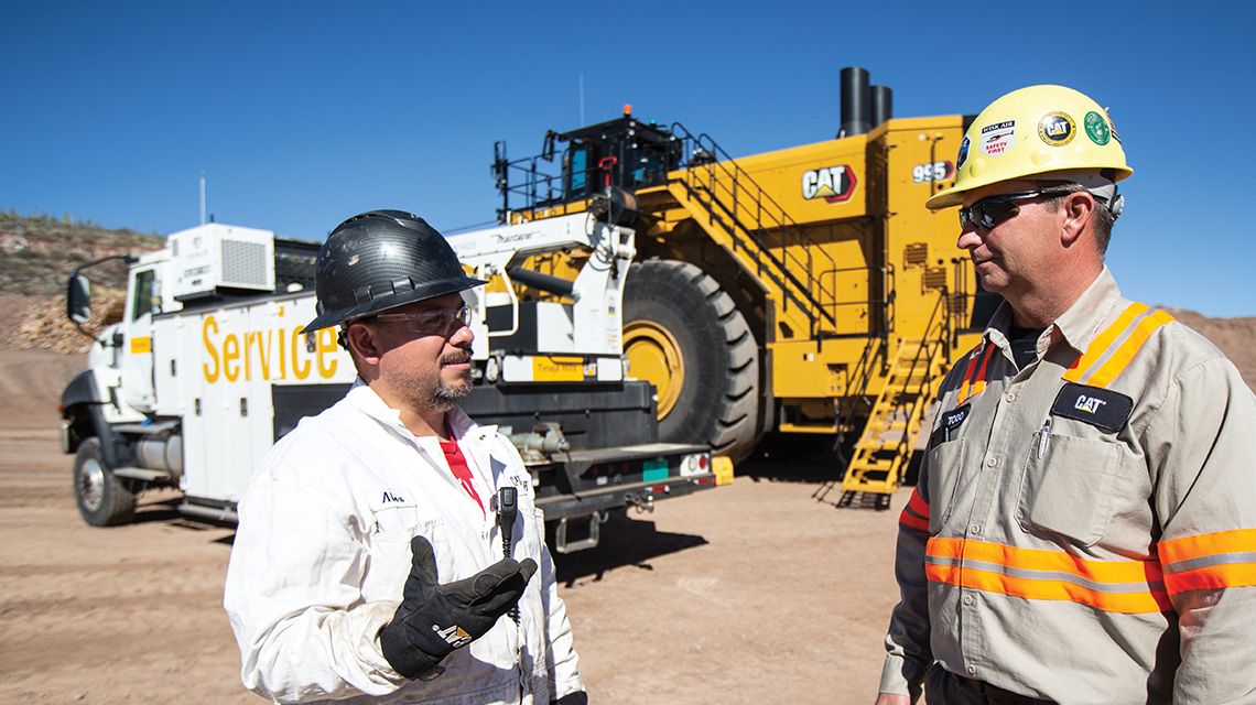 Dos hombres hablando cerca de un camión de servicio estacionado junto a un camión para la minería grande