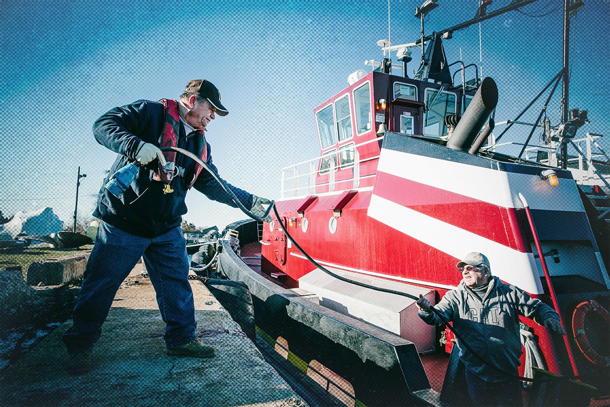Two men in work clothing and ball caps are feeding a hose aboard a red and white tugboat under a blue sky.