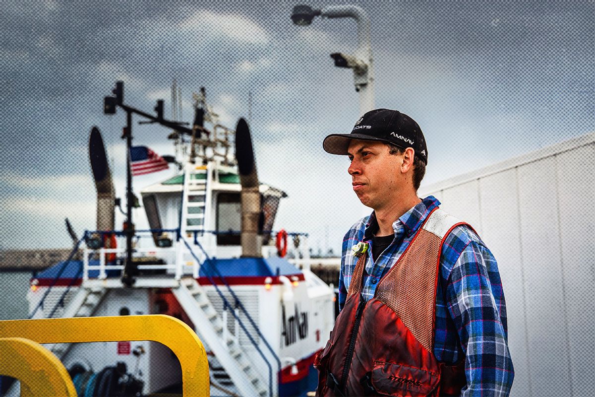 Man wearing a dirty, orange mesh-shouldered life jacket over a blue flannel shirt stands in front of a docked tugboat flying an American flag.