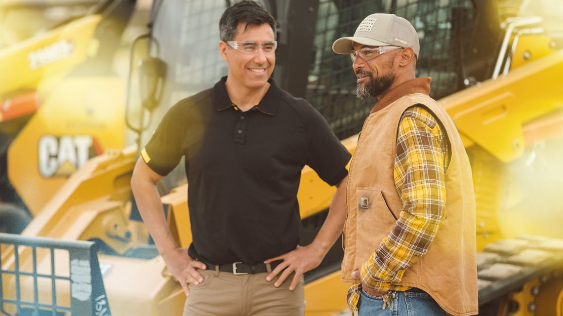 2 men smiling and talking in front of an excavator