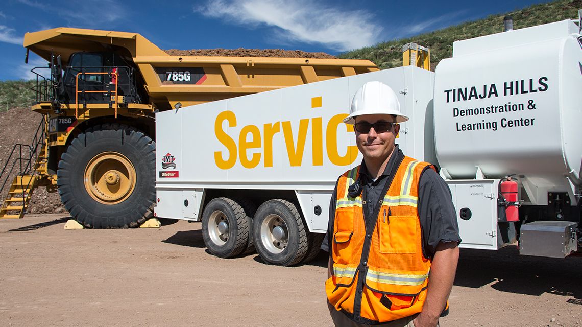 Man smirking wearing white helpmet andorange high-vis vest. Service truck and mining truck in background