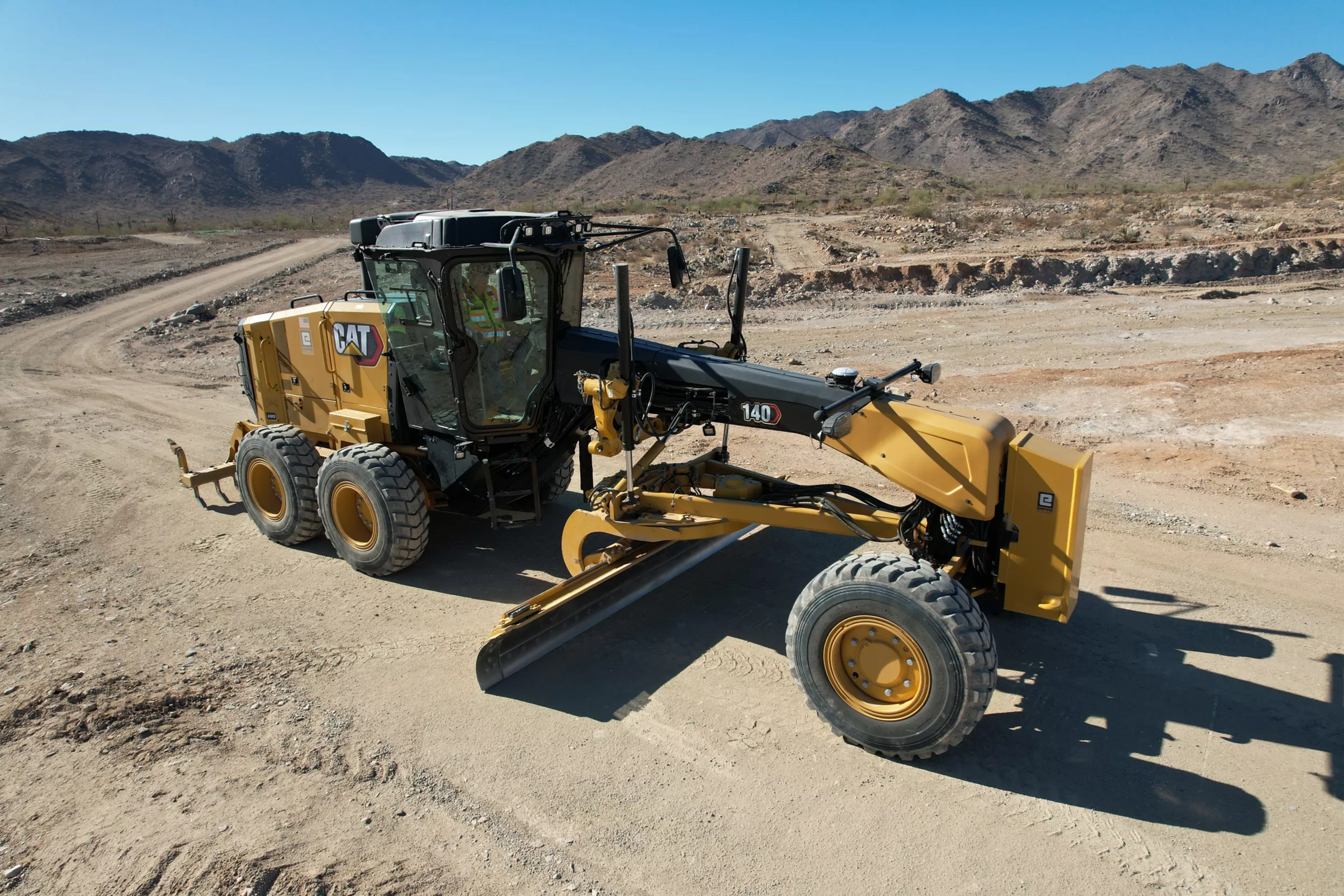 140 Motor Grader in the desert. The machine has the center moldboard resting on the ground and the machine is not in motion. There is a rear ripper, a front counterweight, and a Joystick control configuration.