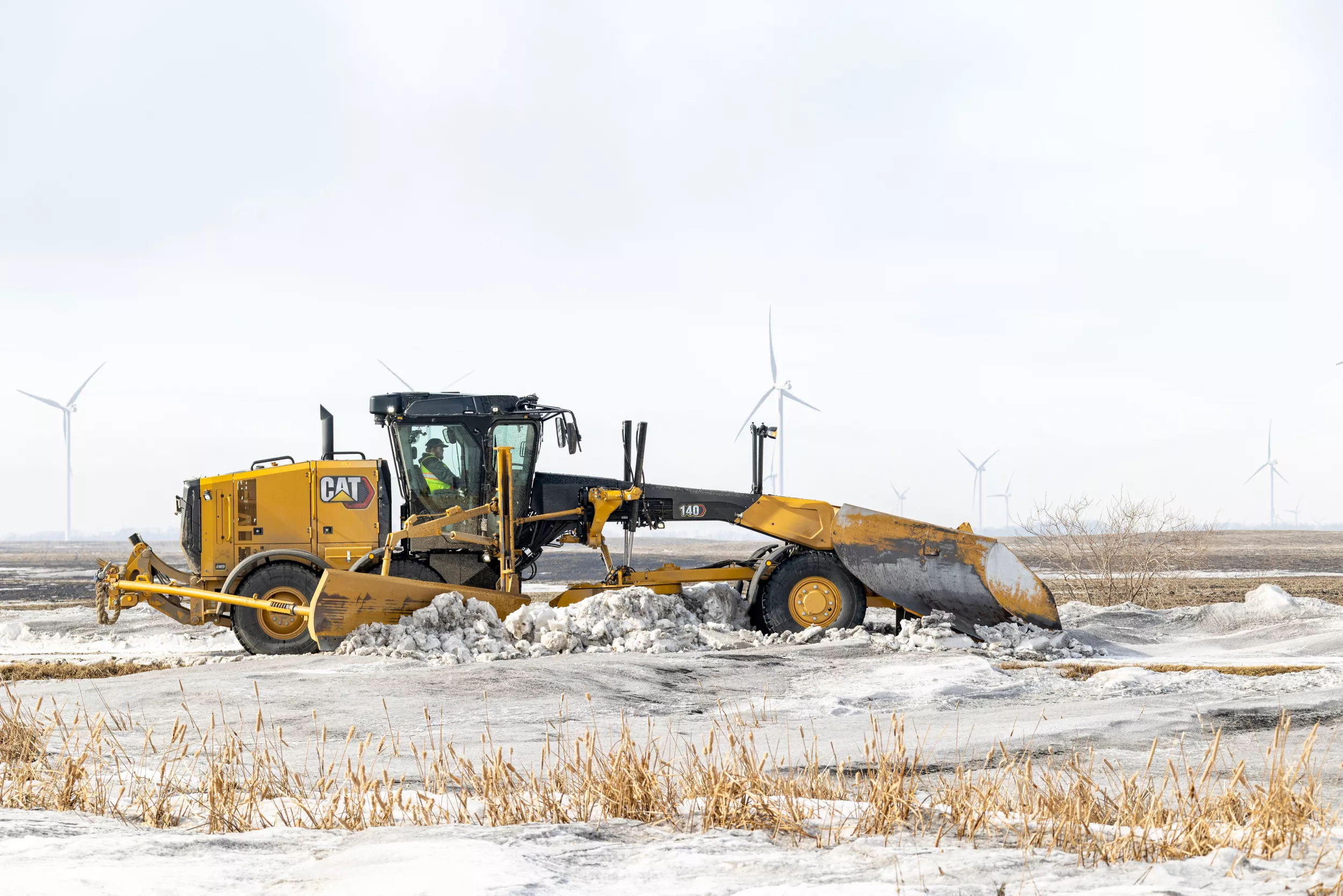 140 Motor Grader pushing snow with a snow wing on the side and a v-plow on front.