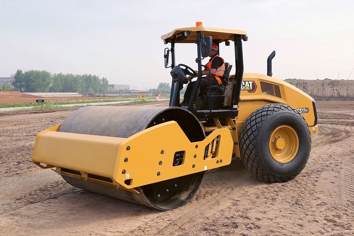 Un hombre en una compactadora de suelo vibratoria Caterpillar CS11 en un campo de tierra.