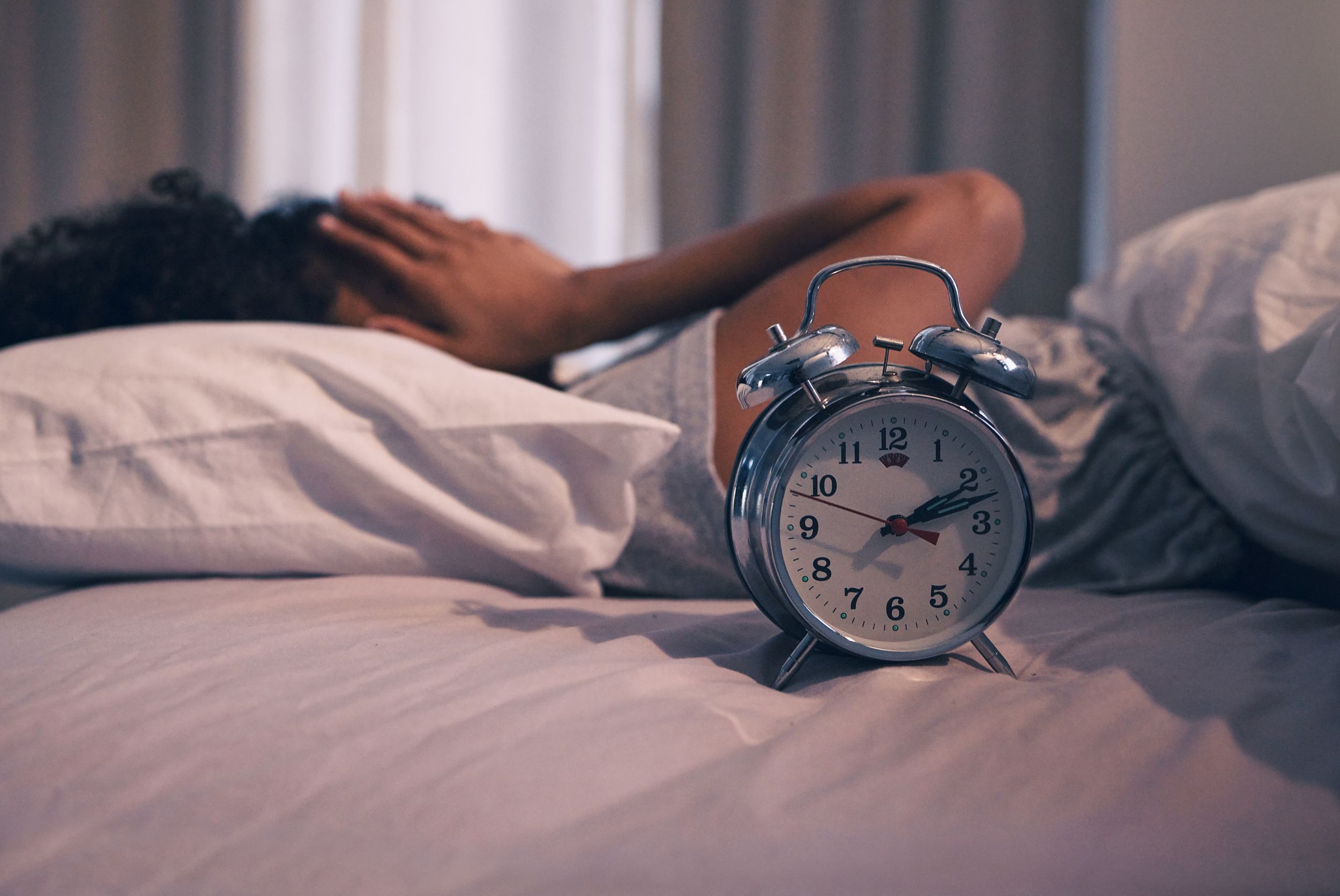 Exhausted woman trying to sleep with alarm clock sitting next to her on bed