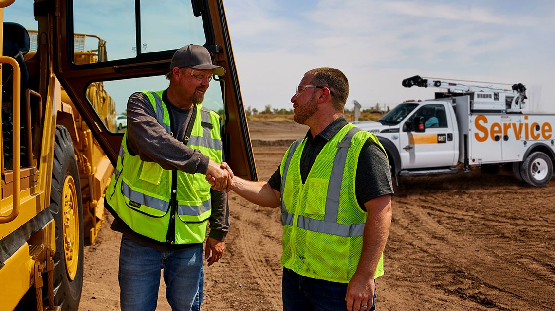 two men in safety vests shake hands with a Cat Service vehicle in the back ground