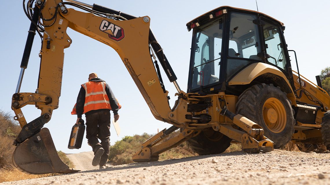 A heavy machine operator carries a seals and fasteners kit near a Cat 420 awaiting maintenance. 