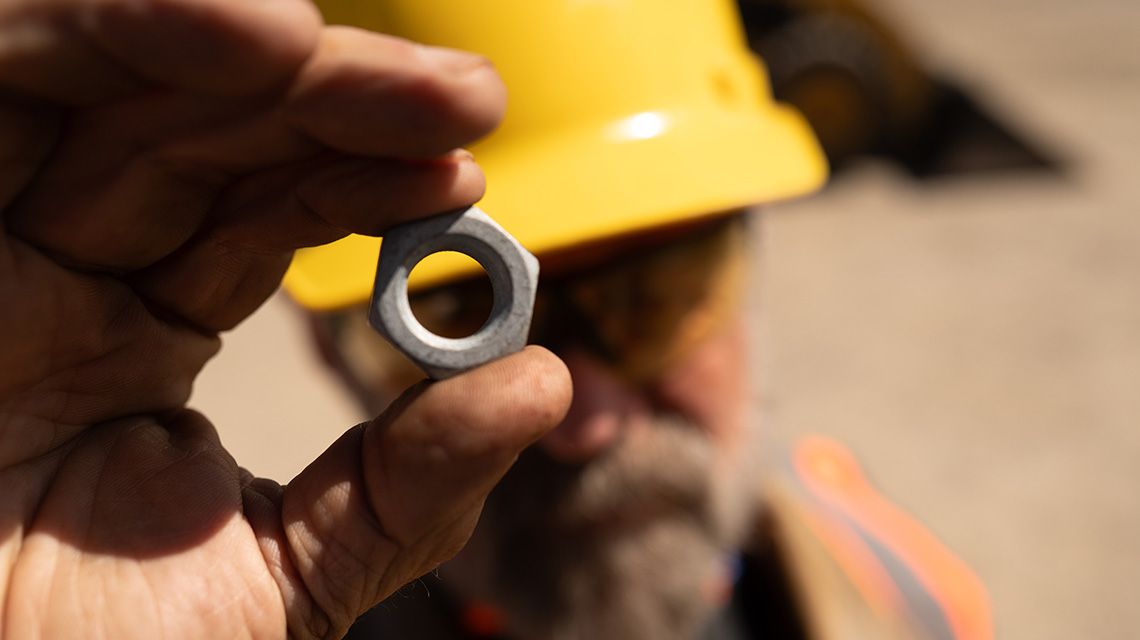 A close-up of a blurred construction professional holding a metal nut part with a yellow hard hat.