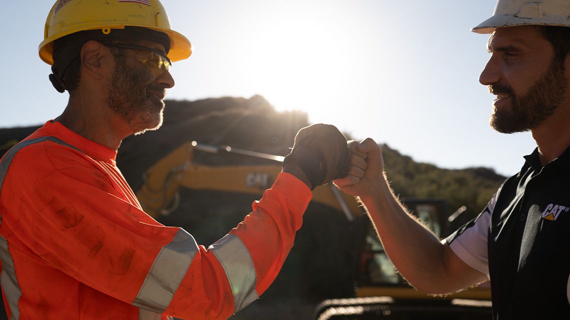 A heavy machine operator and his Cat dealer fist bump on a sunny day with an excavator in the background.