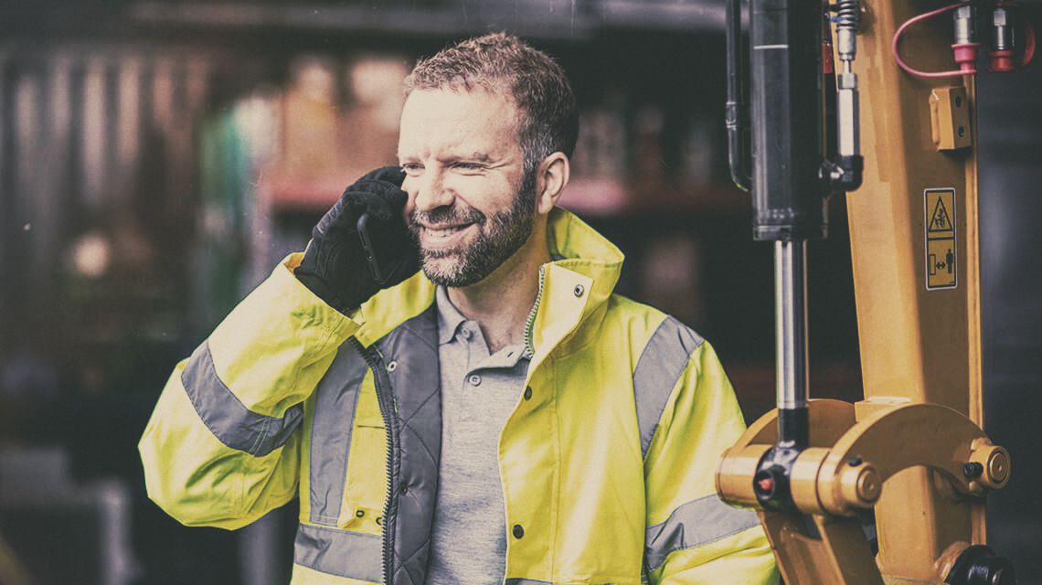 Man in a high-visibility jacket talks on a phone beside construction equipment.