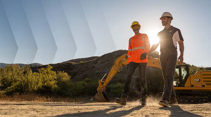 Construction professionals wearing appropriate safety gear walk across a dusty job site with a yellow Caterpillar excavator in the background at sunset.