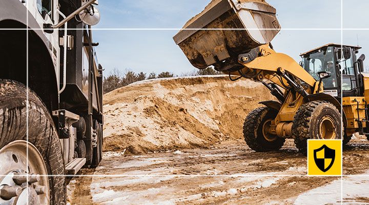 A Cat wheel loader operating on a rugged construction site, loading material next to a commercial dump truck during winter maintenance work. 