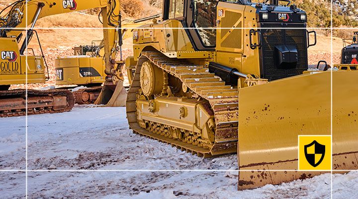Detailed view of a Cat dozer undercarriage, tracks, and blade on snowy ground, featuring a yellow and black shield protection logo 