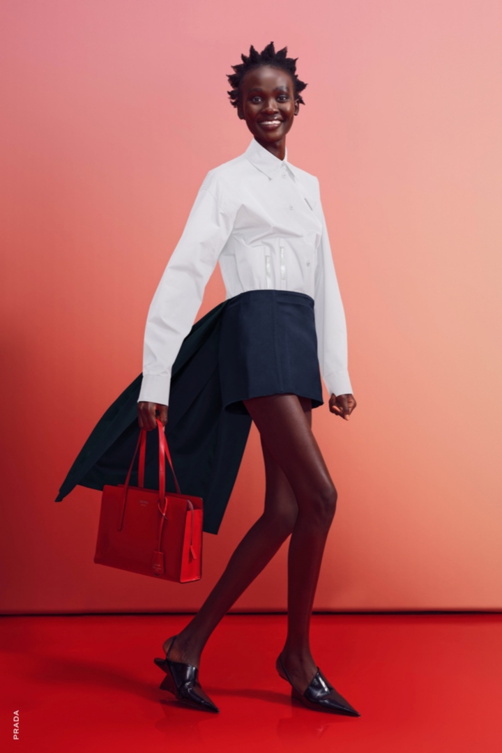 A female model wearing a shirt, skirt, and pointy toe pumps and carrying a bag.