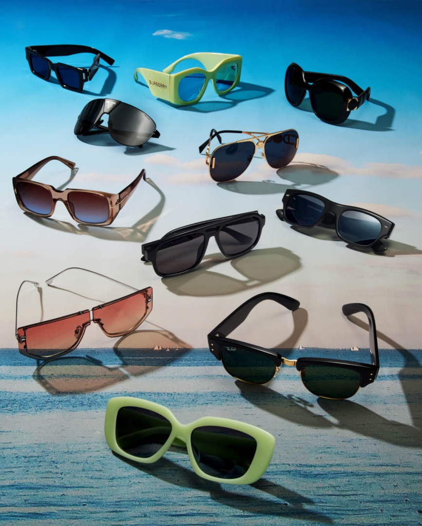 Un assortiment de lunettes de soleil de formes, de tailles et de couleurs différentes devant un arrière-plan montrant un ciel bleu au-dessus d’un océan. 