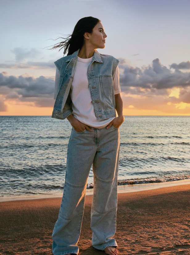 Ani Wells wearing a denim vest and jeans, posing on a beach at sunrise.
