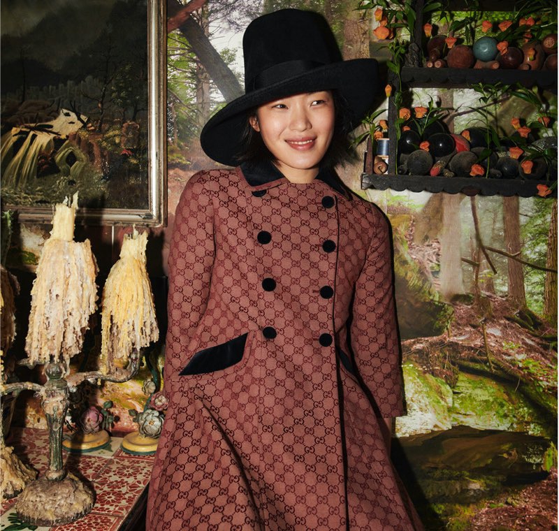 A grinning model in a double-breasted dress coat with an allover GG pattern leans against a table with dripping candles.