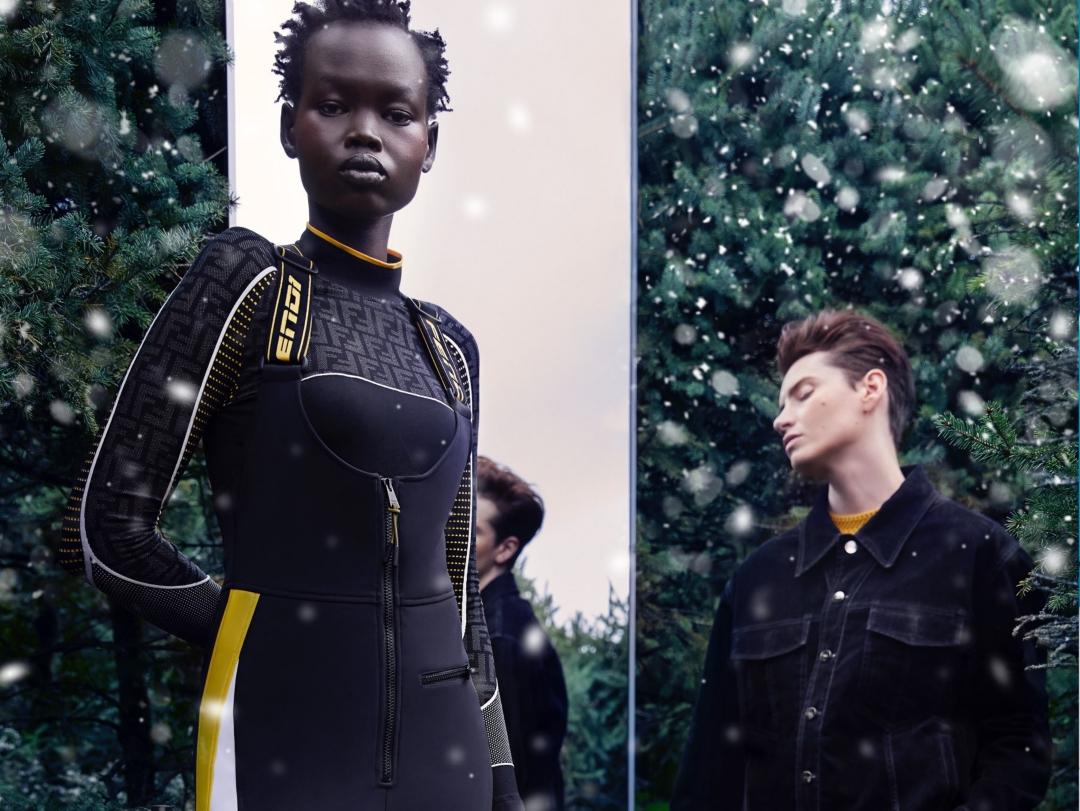 Two models standing in front of a mirror and a hedge—the female model wearing a Fendi ski jumpsuit in black and yellow, and the male model wearing a black outfit.