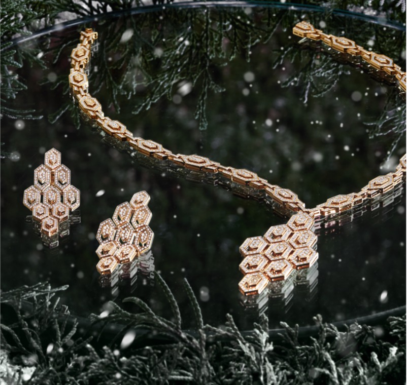 Paire de boucles d’oreilles en or avec diamants et collier de diamants, déposés sur une surface réfléchissante et entourés de feuillage vert festif.