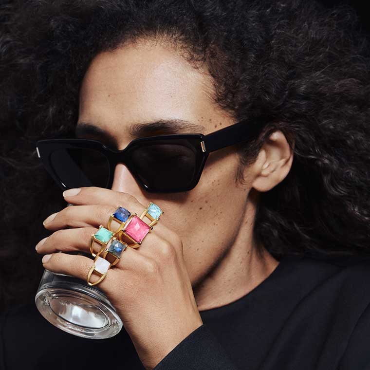A model wearing black sunglasses and colourful rings while drinking from a glass.