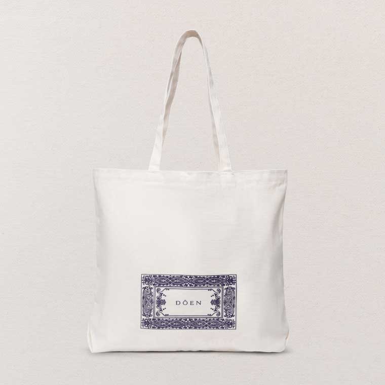 A white tote bag with a navy patterned Doên logo on the front of it.