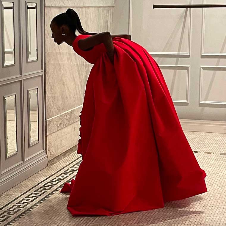A model wearing a red dress, bending at the waist to read a sign on a wall.