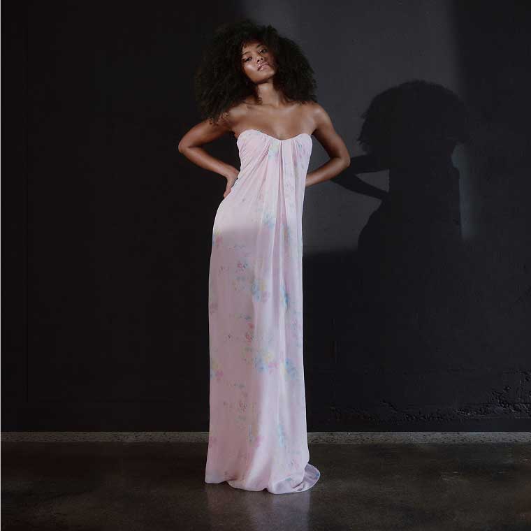 A model wearing a long strapless pink gown posing in front of a dark backdrop.
