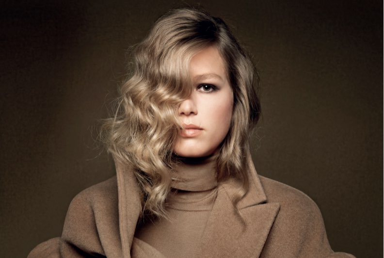 A blonde model wearing a camel coloured coat and turtleneck against a camel coloured backdrop