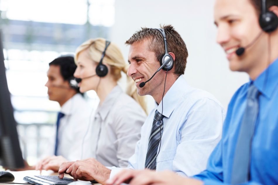 Friendly representative smiling while assisting someone over the phone, with colleagues on either side - copyspace