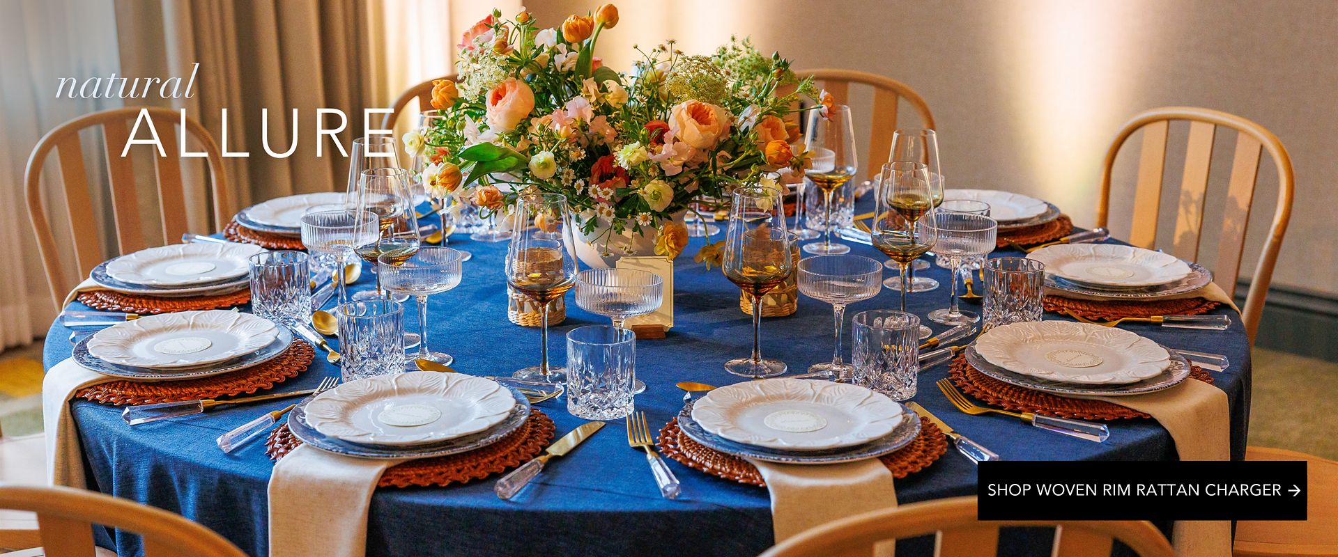 Elegant dining table styled with blue linen, woven rattan charger plates, crystal glassware, and lush peach-and-white floral centerpieces