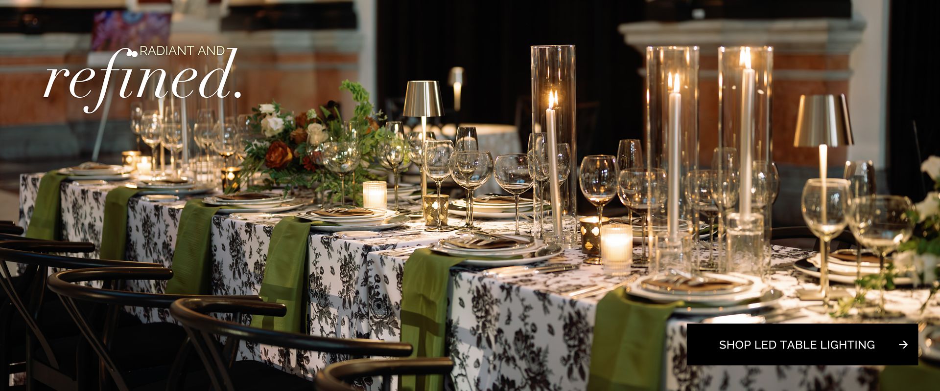 An elegant dining table set for a formal event, featuring a black and white floral tablecloth, olive green napkins, and sophisticated LED table lamps for rent