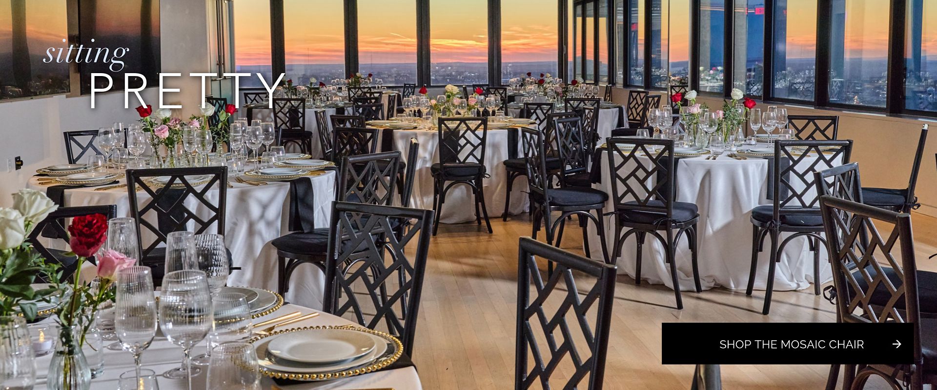 Black Mosaic Chairs with cushions at white-linen tables with gold chargers and roses in a sunset-view event space