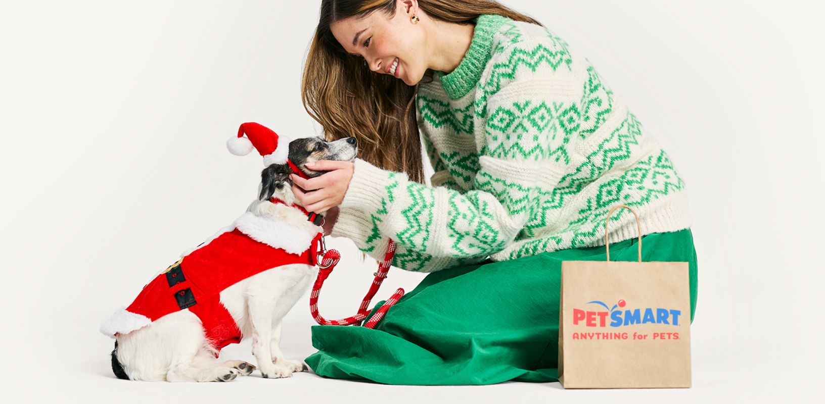 Woman kneeling down next to a PetSmart shopping bag and petting her dog who is dressed in a Santa costume