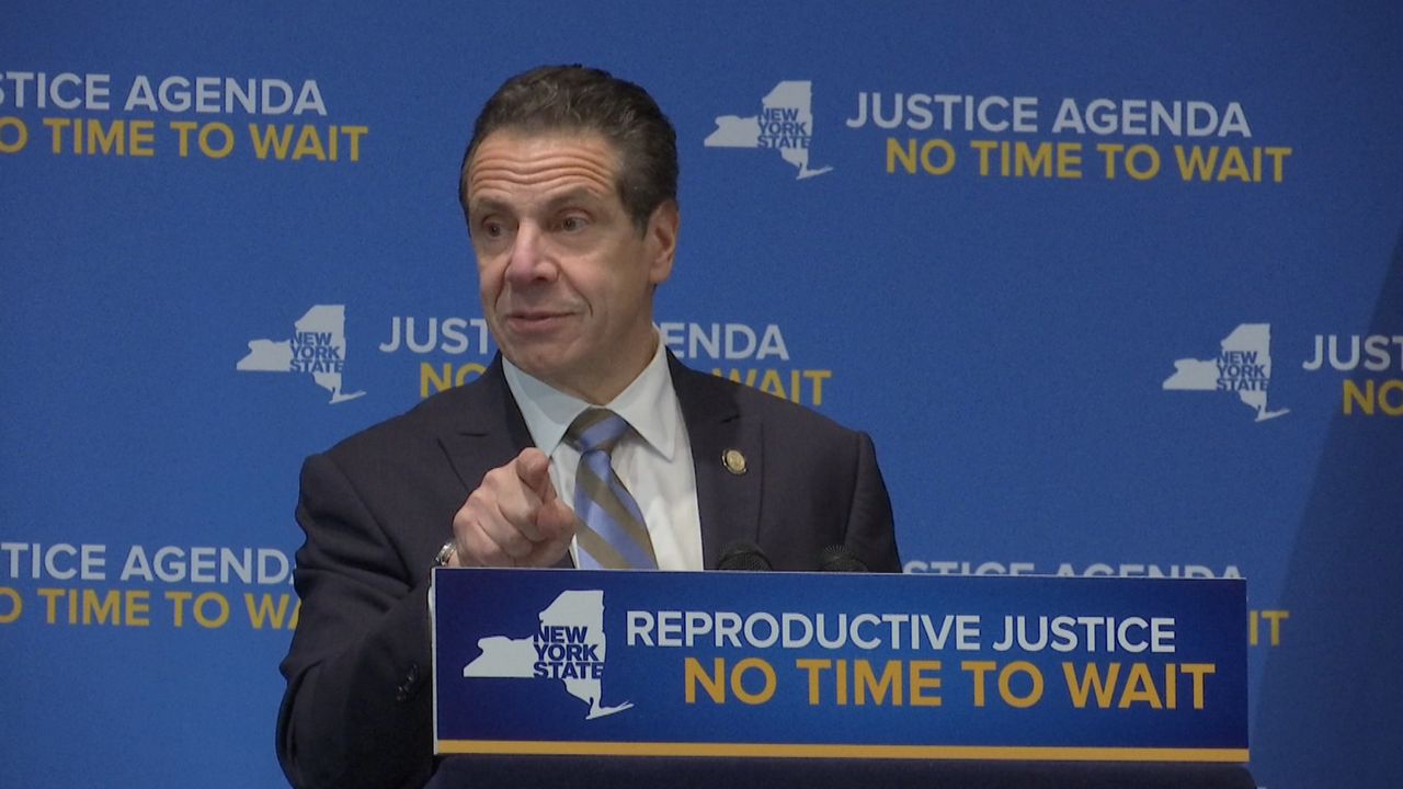 New York Gov. Andrew Cuomo, wearing a black suit jacket, a white dress shirt, and a blue tie, stands in front of a blue sign and blue wallpaper that reads, in white and yellow text, "Justice agenda no time to wait."
