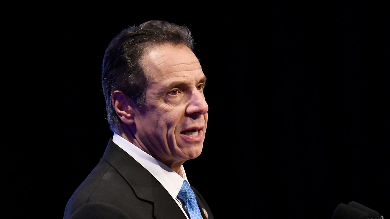 New York Gov. Andrew Cuomo, wearing a black suit jacket, a white dress shirt, and a blue tie, speaks into two black microphones while delivering his State of the State address at the Empire State Plaza Convention Center in Albany.