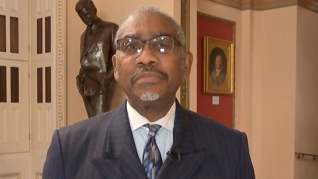 Queens Rep. Gregory Meeks, wearing square glasses, a black suit jacket, a white dress shirt, and a black tie, stands in a large hall on Capitol Hill.